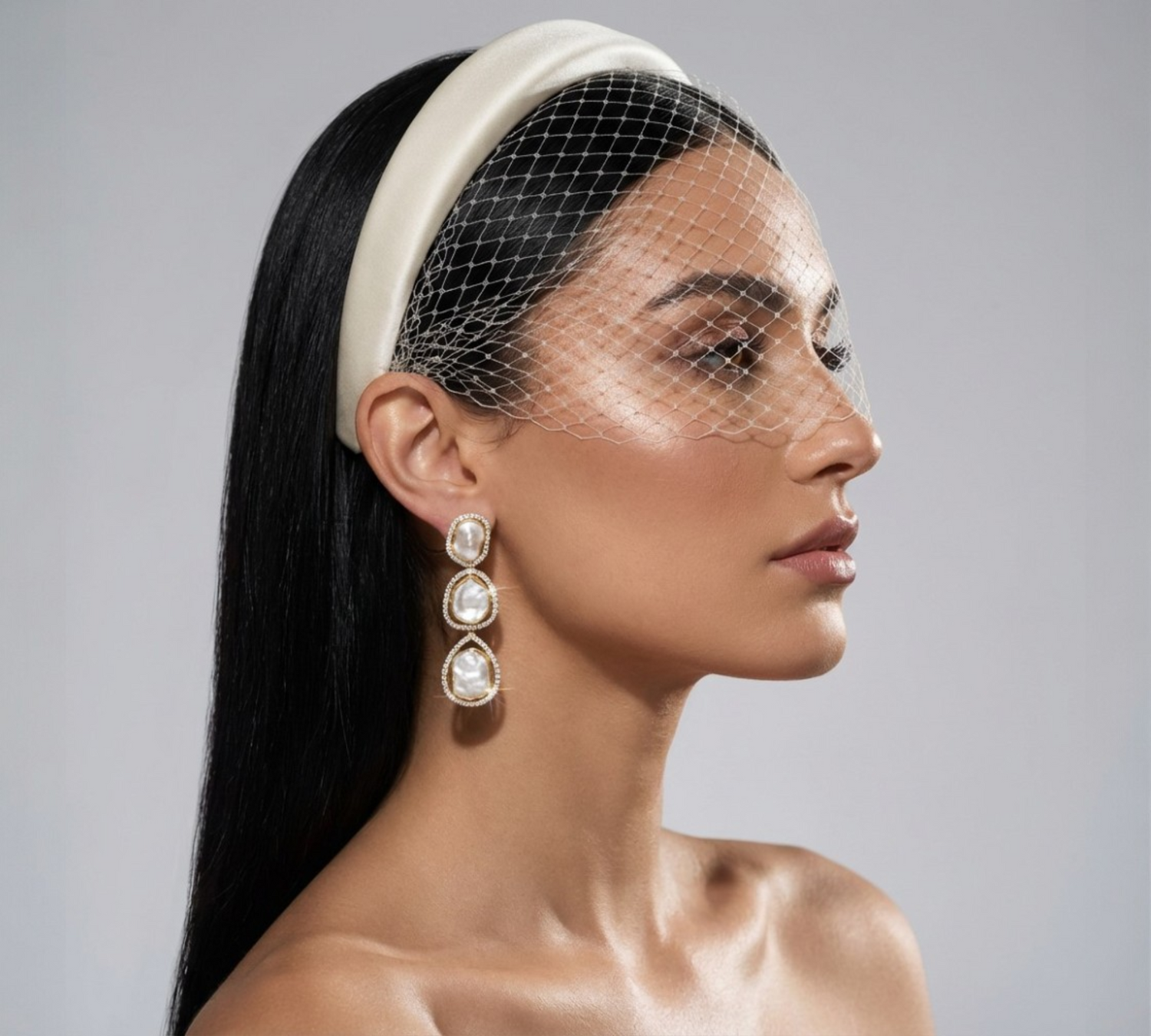 High-fashion bridal portrait featuring pearl drop earrings, satin headband and birdcage veil in a minimal studio setting.