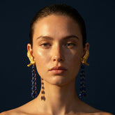 Woman wearing gold earrings with blue beads against a dark background. Celestine Flight lapis lazuli statement earrings with sculptural gold birds by Tania Claire Australia